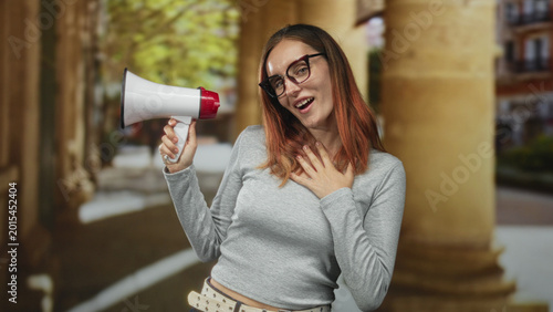 Bild auf Leinwand Woman holding megaphone with hand on chest in a stone building colonnade, wearing glasses and grey top; sincere appeal
