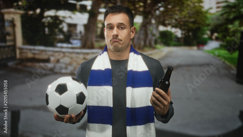 Photography Hispanic man holding soccer ball and beer bottle on a residential street, blue white scarf over shoulders, pouting and shrugging with closed eyes; disappointment solitude