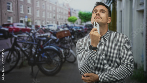 Photography Young hispanic man holding smartphone near chin while looking up on a city street; thoughtful planning