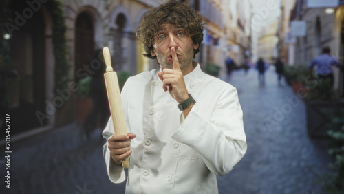 Фотография Man chef in white uniform holds a rolling pin and places finger to lips for silence on a narrow cobblestone street with blurred passersby; playful secrecy