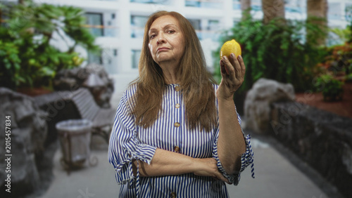 Valokuva Senior woman holds a lemon aloft with raised hand and crossed arms, neutral stern gaze toward camera in building; contemplation patience resolve