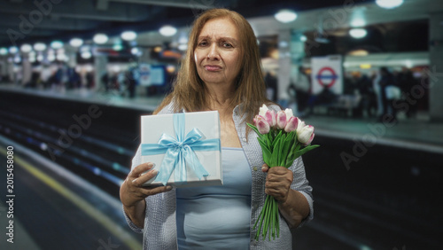 Foto Woman holding wrapped gift box and clutching a bouquet of pink tulips, hands visible and frowning on a train station building platform; waiting delay sadness