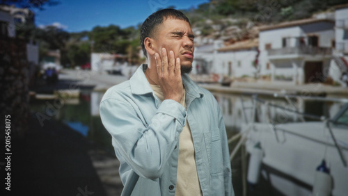 Photography Young hispanic man with hand on cheek wincing beside a docked sailboat in front of white seaside building; pain