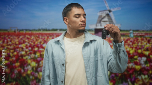 Man holding car keys in raised hand near a windmill building in a tulip field, looking at key; quiet contemplation.