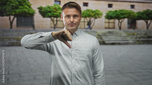 Cuadro en lienzo Man showing thumbs down gesture in front of a building on stone steps and plaza beside trimmed trees and cobbled paving; disapproval