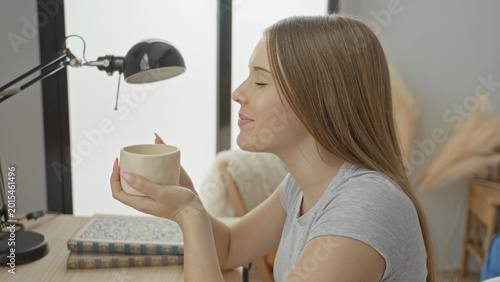 фотографія Young woman sitting at desk holding ceramic mug and inhaling coffee aroma near lamp and books in studio; calm morning pause