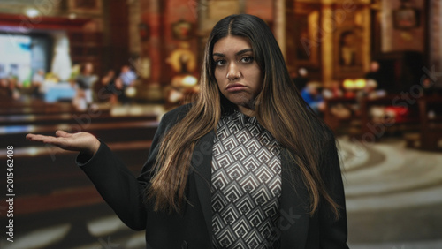 Фотография Woman in blazer and patterned top shrugging with palm up and sullen pout amid ornate interior of a historic building; quiet resignation