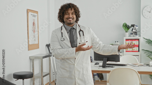 Photos Man doctor in white coat with stethoscope, smiling and gesturing with open hands during consultation in clinic building; friendly reassurance