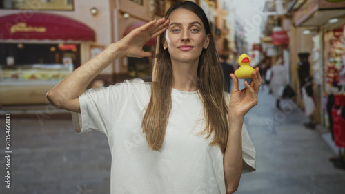 Fotografía Woman holding a yellow rubber duck and saluting with hand to forehead on a busy street market; playful innocence