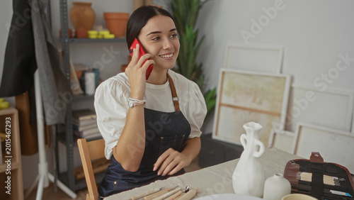 Tableau sur toile Woman in apron holds red phone to ear and gestures with open hand, seated at work table with ceramic vases and sculpting tools in pottery studio; cheerful conversation