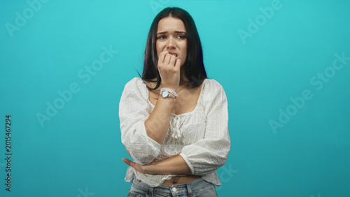 Valokuva Woman biting fingernails and holding hand to mouth in studio with teal backdrop, white blouse and jeans; anxiety uncertainty
