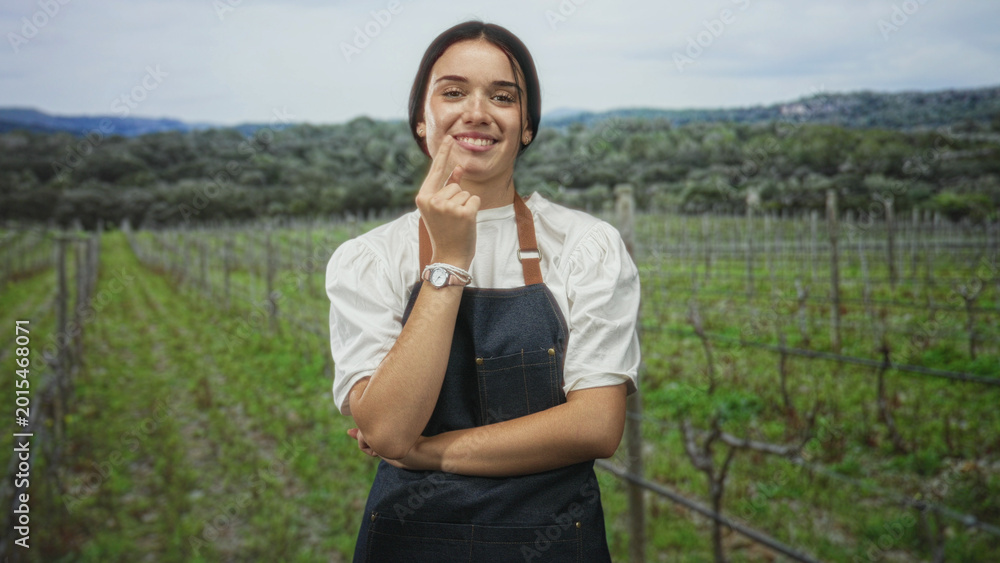 Obraz premium Woman in apron touches chin with hand, arms folded, standing among vineyard rows in forest setting with distant hills and olive trees; calm thinking.