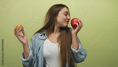Foto Woman holding a red apple and a cupcake in a light green studio, glancing at the apple while comparing choices; choice indecision