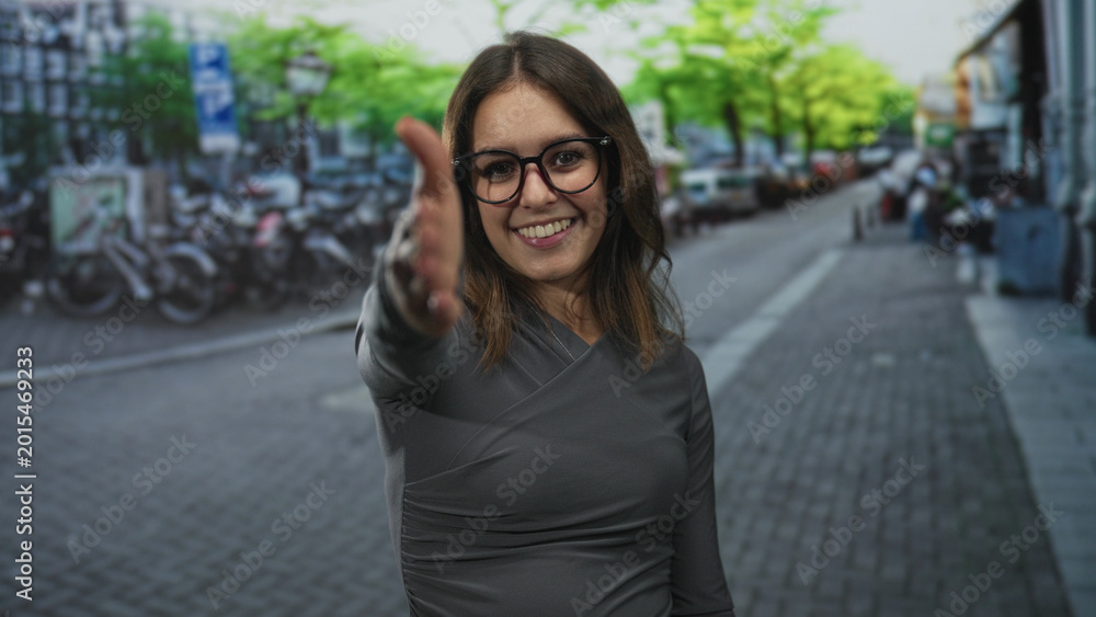 Fototapeta premium Young hispanic woman extends hand for handshake on city street, smiling and wearing glasses and grey top; friendly greeting.