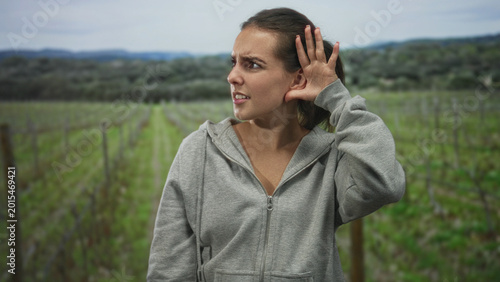 Woman with hand to ear for listening in a forest field vineyard wearing gray hoodie and frowning; annoyance.