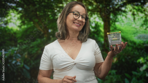 Foto Woman holding a miniature shopping cart in hand, smiling and posing in a forest setting; cheerful thrift savings