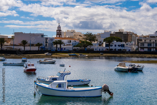 Vibrant scene of Arrecife Lagoon in Lanzarote featuring numerous boats floating peacefully in clear waters. Modern buildings and distant mountains complete the tranquil backdrop