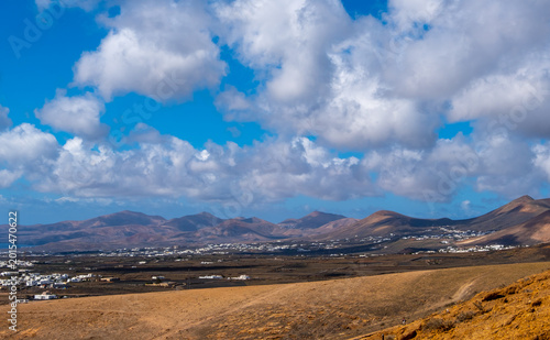 Scenic view of a Lanzarote village nestled at the foot of a volcanic hill under a dramatic cloudy sky. The contrast of white buildings against the rugged terrain highlights Lanzarote unique landscape