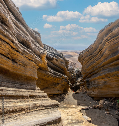 Rugged canyon landscape of Lanzarote with striking layered rock formations under a bright blue sky. The geological formations emphasize the island's volcanic origins