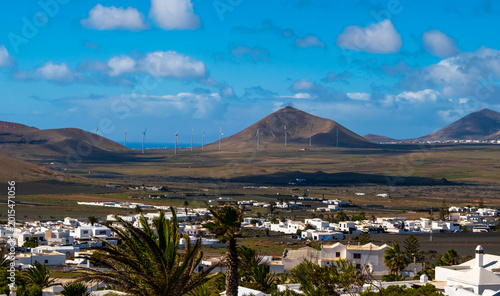 Scenic view of a Lanzarote village nestled at the foot of a volcanic hill under a dramatic cloudy sky. The contrast of white buildings against the rugged terrain highlights Lanzarote unique landscape