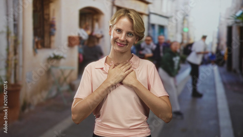Foto Woman blond with hands over heart wearing pink shirt, smiling softly amid blurred pedestrians on a narrow street; quiet gratitude