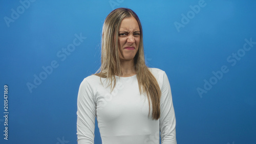 Billede på lærred Woman scrunches face and wrinkles nose while standing in studio set with vibrant blue backdrop; disgust aversion