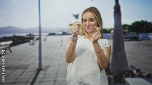 Tableau sur toile Woman makes finger heart gesture with hands on seaside promenade wearing white blouse and smartwatch, palms visible; playful flirtation
