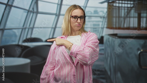 Photography Woman points index finger left in airport lounge, wearing pink striped shirt and glasses; disapproval assertiveness