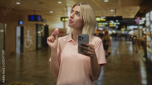 Konstfotografi Woman holding pink pastry and smartphone while checking messages and glancing at the screen in a busy airport concourse; curiosity leisure