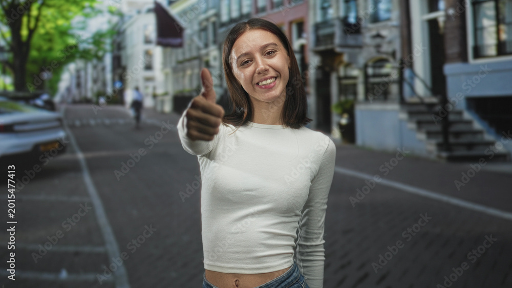 Fototapeta premium Young hispanic woman smiling and giving thumbs up with bare thumb visible on a street lined with buildings; confidence joy.
