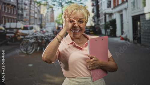 Photos Woman holding pink clipboard grips folder and makes ok sign to eye with her hand on a street near bicycles and shops; joyful confidence