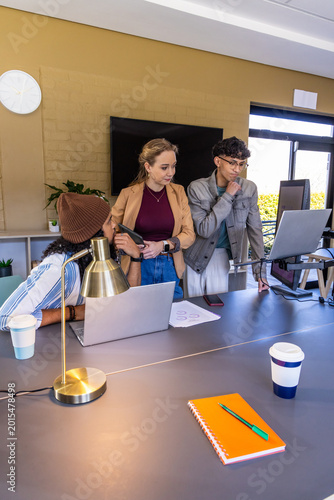 Diverse coworkers standing and leaning around conference table, holding smartphone, using laptops