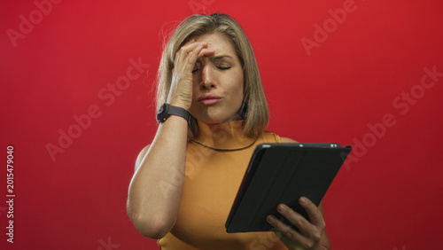 Schilderij op canvas Blonde young woman holding tablet, hand to forehead and raised palm while reading screen in studio with red wall; surprise