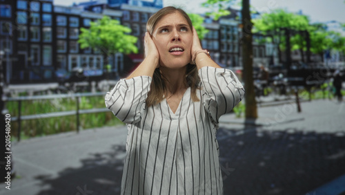 Photos Woman in striped shirt covers ears on busy street beside canal railings under bright sunny sky; annoyance