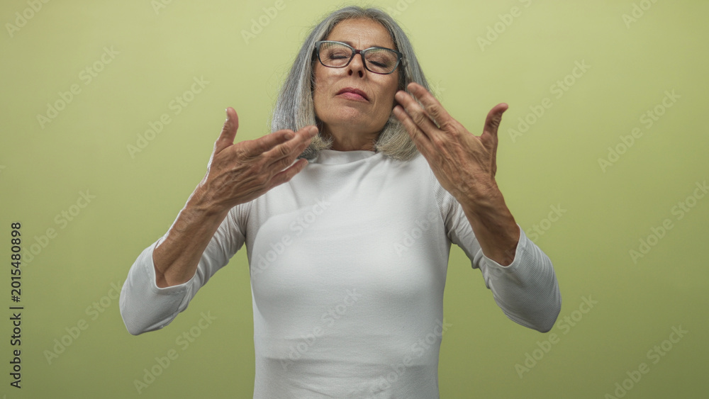 Naklejka premium Middle aged grey haired woman with hands on chest and glasses in studio green backdrop; self care serenity.