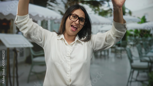 Obraz na płótnie Woman, young hispanic, wearing glasses, arms raised and open mouth gesture in building terrace outdoors near cafe seating; playful fun energy