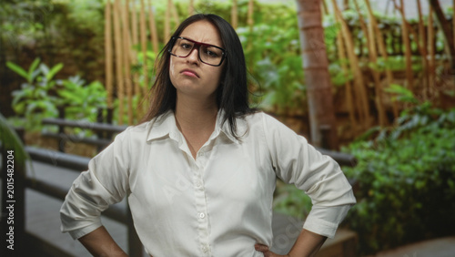 Foto Woman wearing glasses and a white shirt, hands on hips on a wooden forest walkway flanked by green plants, stern expression; disapproval