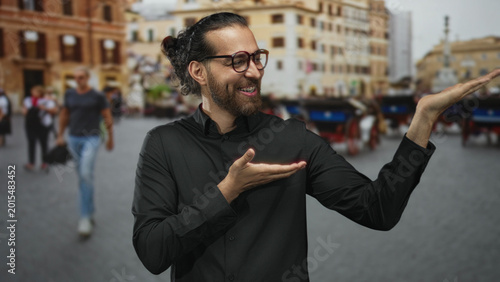 Fényképezés Man wearing glasses and black shirt smiling with palms up on a busy city street plaza beside horse carriages and historic buildings; joy travel welcome
