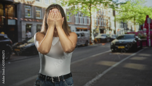 Принти на полотні Woman in white tank top and jeans with hands covering face on busy urban street with parked cars and sidewalk cafe; surprise playful curiosity