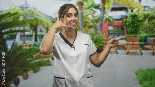 Quadro su tela Woman beautician shows call me hand gesture with left hand and points right index finger by resort pool lounge chairs in building courtyard; playful service