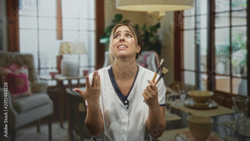 Bild auf Leinwand Young blonde woman wearing white uniform holding brushes, hand raised and gesturing in a building living room with sofa, lamp and table visible; frustration