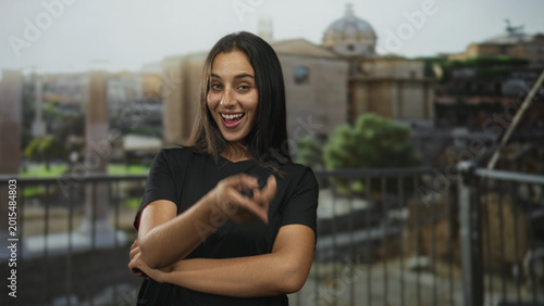 Foto Hispanic young woman with crossed arms and a smile points finger to face on a building terrace overlooking roman ruins; confidence