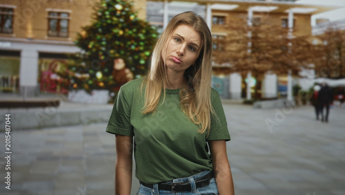 Canvas Print Woman pouting lips, head tilted, wearing green t shirt and jeans on a street by a decorated christmas tree; holiday sadness