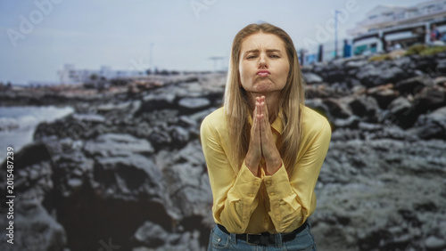Photos Woman pressing hands together in prayer with pursed lips, wearing yellow shirt and jeans in a studio rocky coast backdrop; pleading hope