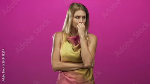 Fotografija Young blonde woman bites fingernails with right hand while left arm crosses torso in pink studio, worried expression and furrowed brow; anxiety