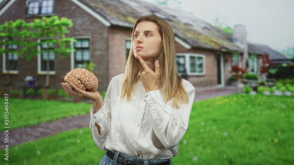 Fototapeta premium Blonde woman holding brain prop, finger on chin while posing in a cottage front yard with green lawn and path; curiosity reflection.