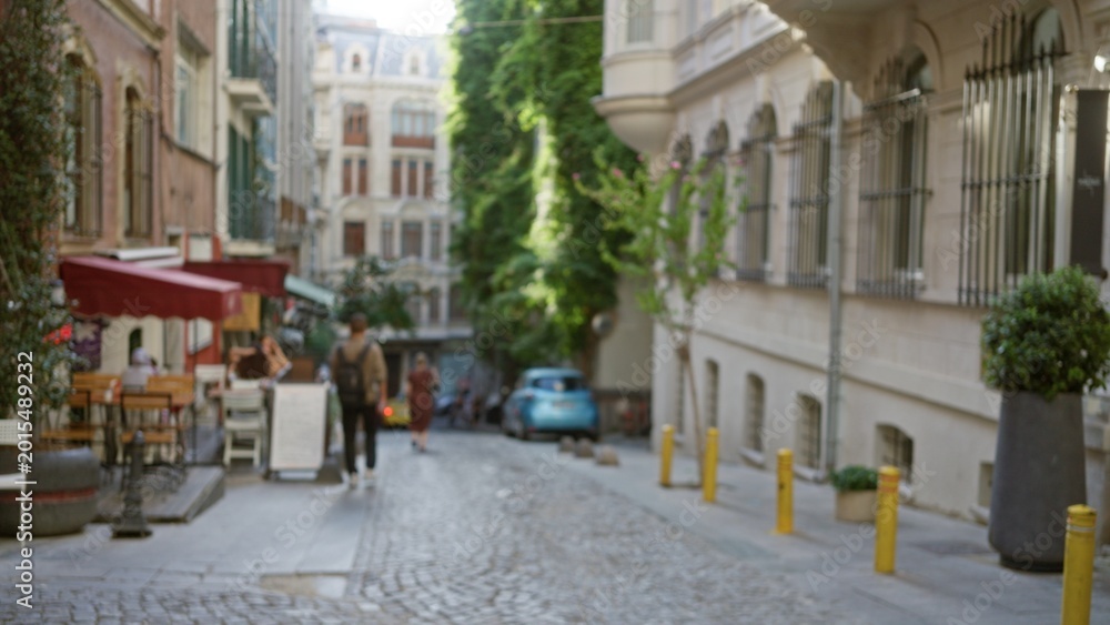 Fototapeta premium Narrow cobblestone street with soft bokeh and defocused shallow focus on storefronts, trees and parked car, outdoor; background copyspace calm.