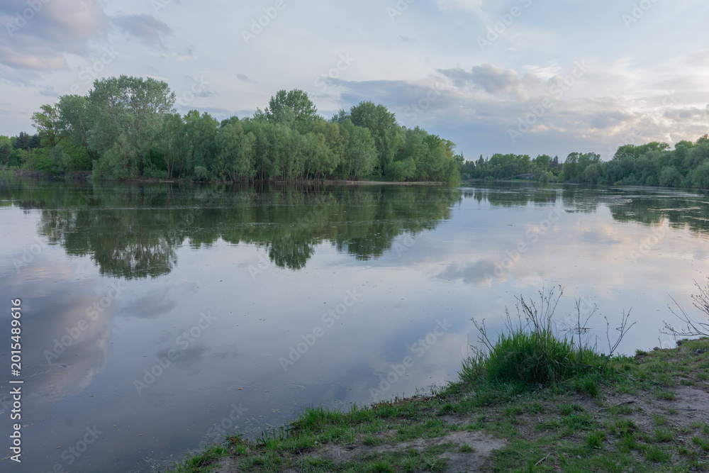 Fototapeta premium a calm river reflecting lush green trees and a cloudy sunset sky.