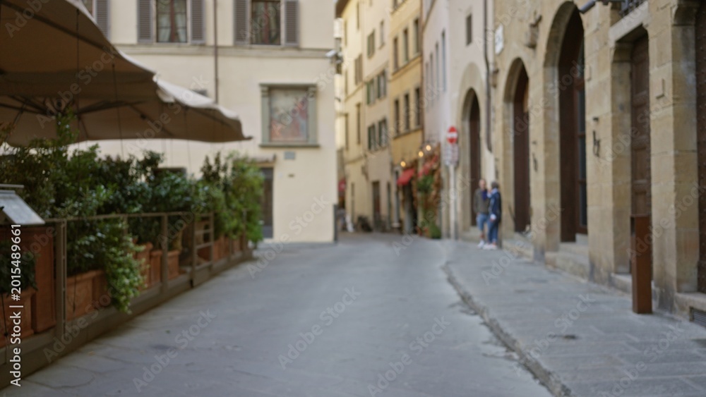 Fototapeta premium Quiet narrow city street in soft defocused bokeh showing blurred stone facades, cafe planters and umbrella, shallow focus outdoor background; background backplate copyspace calm.