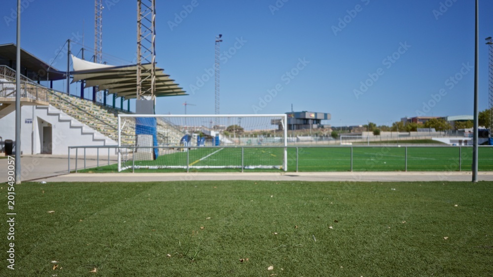 Fototapeta premium Soccer pitch foreground softly blurred with shallow focus on turf and defocused goal and stadium stands visible outdoor; backdrop copyspace backplate template.